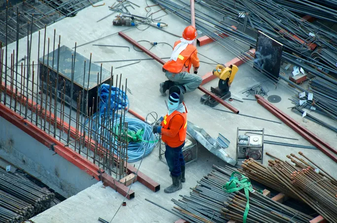 Two construction workers in orange safety vests and hard hats working on a concrete slab surrounded by rebar and tools, seen from above.