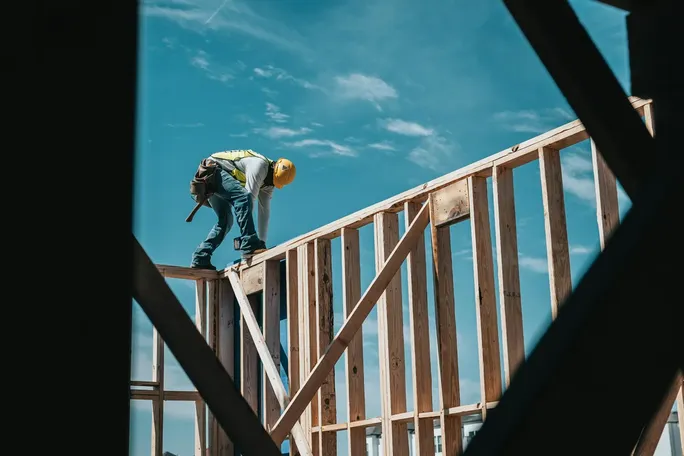 A construction worker in a yellow hard hat and safety vest standing on a wooden house frame against a blue sky.