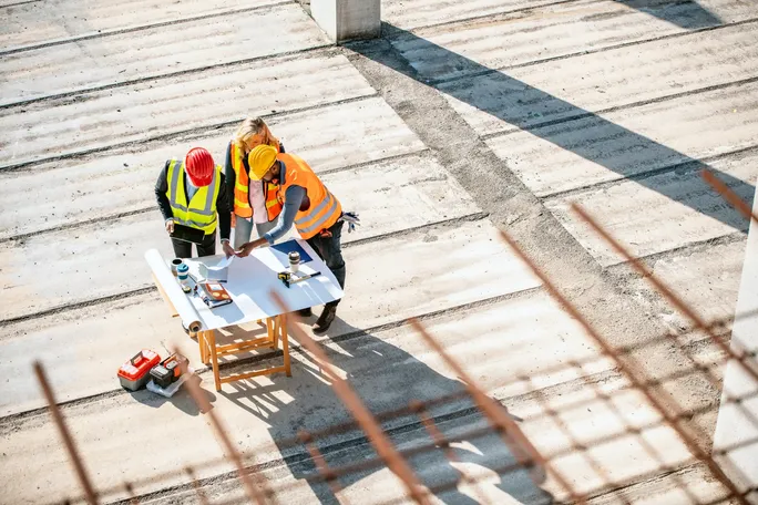 Construction project manager reviewing plans and a tablet with two crew members at a table on a concrete jobsite